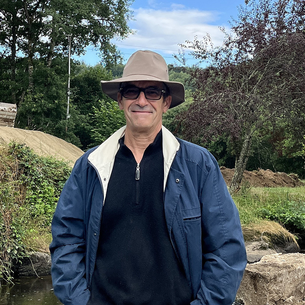 Ein Foto von Marc Bernardot, einem Mann mit Brille und dunklem Anzug, der im Freien steht, mit einer Stadtlandschaft im Hintergrund, darunter Gebäude und ein klarer Himmel. ©Marc Bernardot