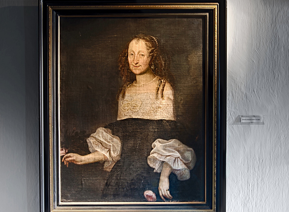 Gemälde einer Frau mit heller Haut und langen, braunen, gelockten Haaren. Sie trägt ein schwarzes Kleid mit weißen, voluminösen Ärmeln und eine Perlenkette. In ihrer linken Hand hält sie eine Rose, eine weitere Rose liegt unten im Bild. Der Hintergrund ist dunkel und neutral. Das Gemälde ist in einem schwarzen Rahmen mit goldenen Verzierungen gefasst und hängt an einer weißen Wand. 