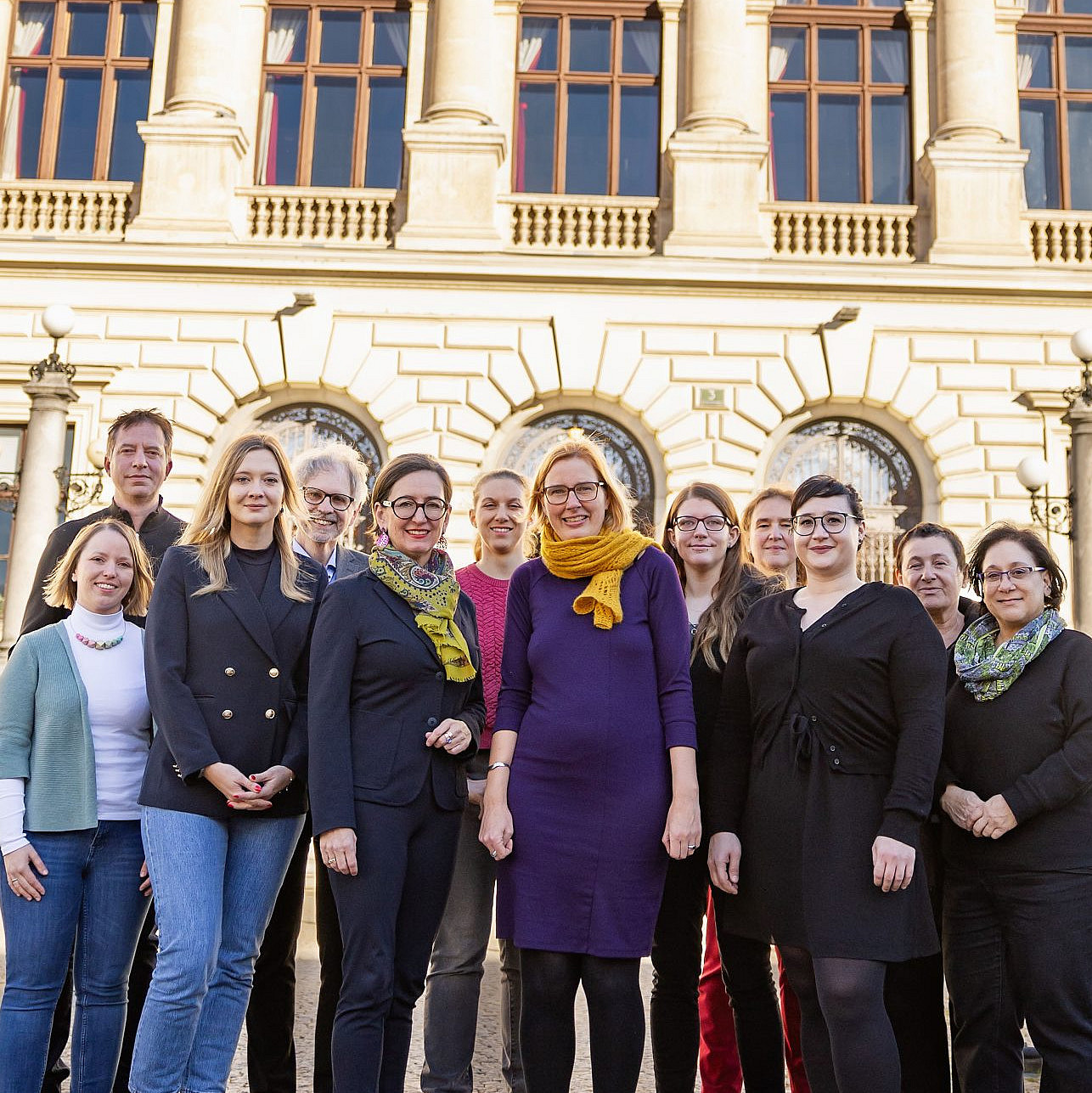 Das Bild zeigt eine Gruppe von Personen, die vor einem beeindruckenden historischen Gebäude mit reicher Fassade und Skulpturen stehen. Sie lächeln und posieren für ein Gruppenfoto. Einige tragen lässige Kleidung, während andere etwas formeller gekleidet sind. Es ist ein sonniger Tag, und das Gebäude im Hintergrund könnte ein Museum oder ein Regierungsgebäude sein. Die Szene strahlt eine fröhliche und lebendige Atmosphäre aus. ©Christine Hofer-Lukic; 15. Österreichischer Zeitgeschichtetag