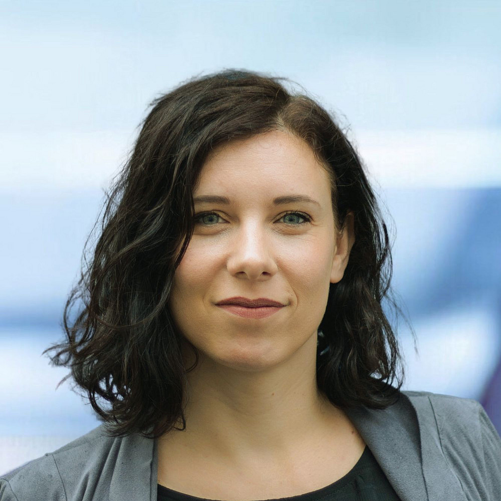 Das Foto zeigt Stefanie Populorum, eine Frau mit welligem dunklem Haar, die ein schwarzes Oberteil und einen grauen Blazer trägt und vor einem unscharfen Hintergrund steht. Sie blickt mit neutralem Gesichtsausdruck direkt in die Kamera. ©Stefanie Populorum