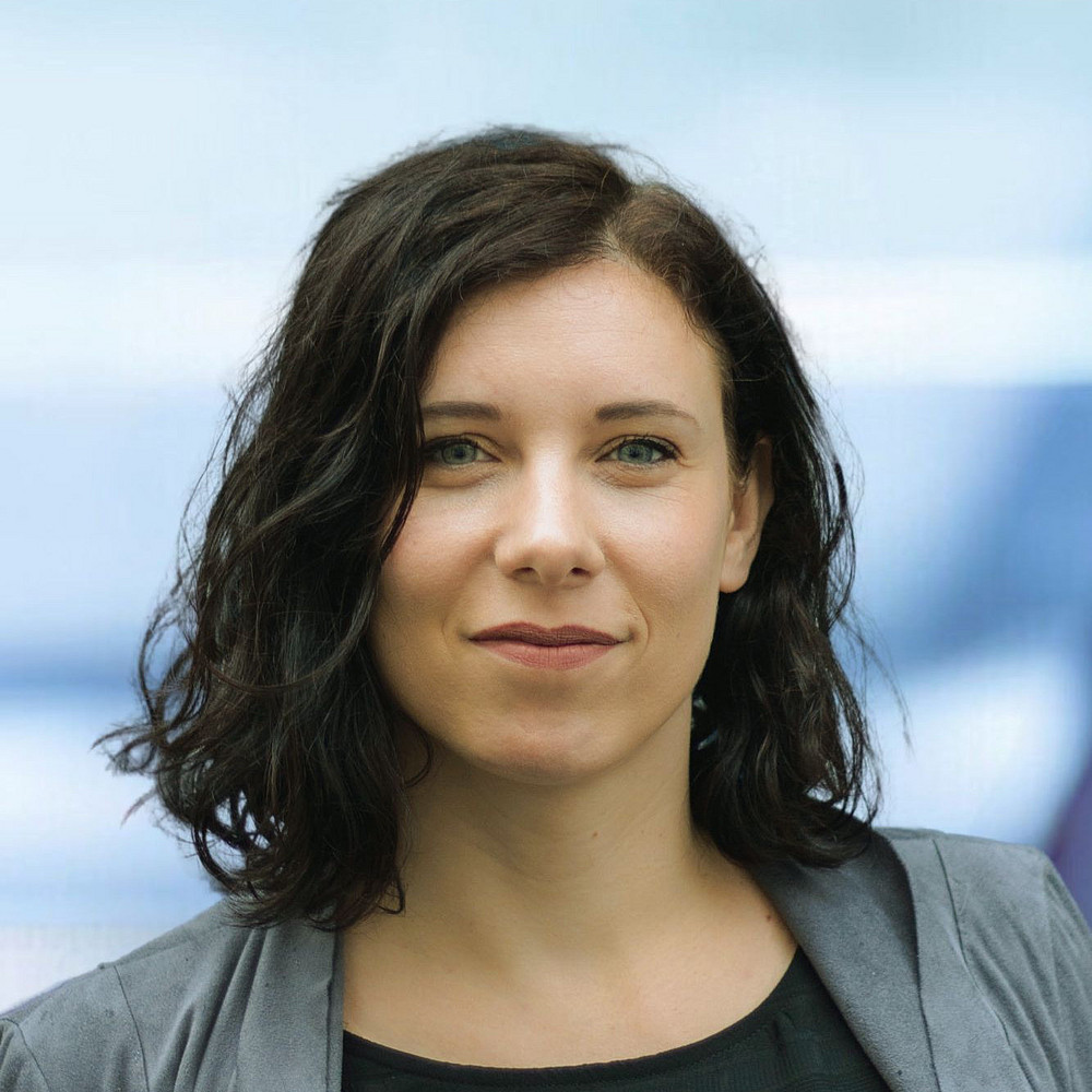 Die Aufnahme zeigt Stefanie Populorum, eine Frau mit dunklen, gewellten Haaren und hellen Augen, die direkt in die Kamera blickt. Sie trägt ein dunkles Oberteil und darüber eine graue Jacke oder einen Blazer. Der Hintergrund ist verschwommen und in hellen Blautönen gehalten, was darauf hindeutet, dass die Aufnahme im Freien oder in einem gut beleuchteten Raum mit einer möglichen Reflexion im Hintergrund stattgefunden hat. Ihr Ausdruck ist neutral bis leicht lächelnd. Die Bildkomposition konzentriert sich auf ihr Gesicht und den oberen Teil ihres Oberkörpers. ©Stefanie Populorum