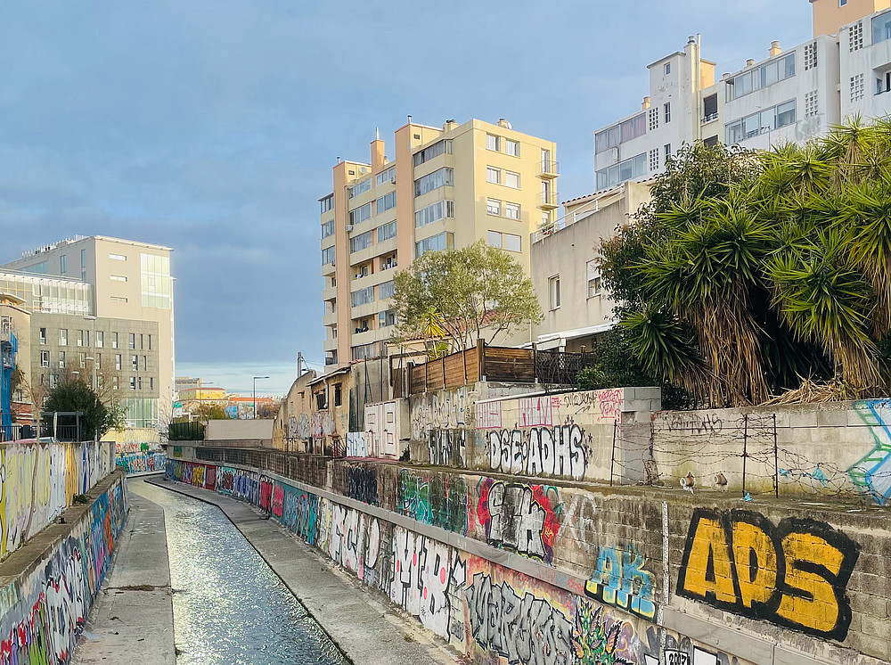 The photo shows a narrow urban waterway whose concrete walls are decorated with colourful graffiti. Tall buildings can be seen in the background under a cloudy sky. Vegetation grows along the water. ©Pierre Sintès
