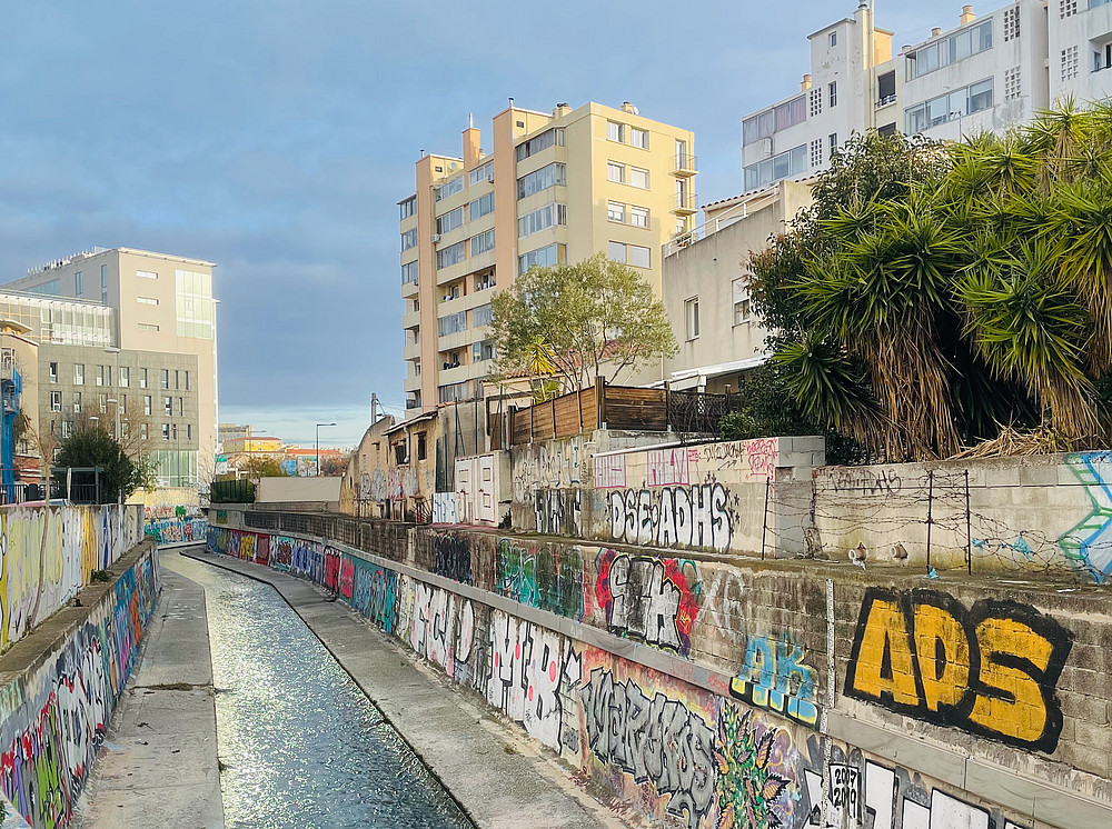 Das Bild zeigt einen Kanal mit Betonwänden und Graffiti in einer städtischen Umgebung, umgeben von Wohngebäuden. Aufgrund fehlender nützlicher Informationen aus der Suche ist es nicht möglich, den genauen Ort oder den konkreten Zeitpunkt mit Sicherheit zu bestimmen. Der Kanal ist mit Beton ausgekleidet und enthält fließendes Wasser. Die Kanalwände sind mit verschiedenen Graffiti und Street-Art-Motiven bedeckt. Im Hintergrund sind Wohngebäude unterschiedlicher Höhe zu sehen. Auf der rechten Seite ist Vegetation, darunter einige Palmen, zu erkennen. ©Pierre Sintès