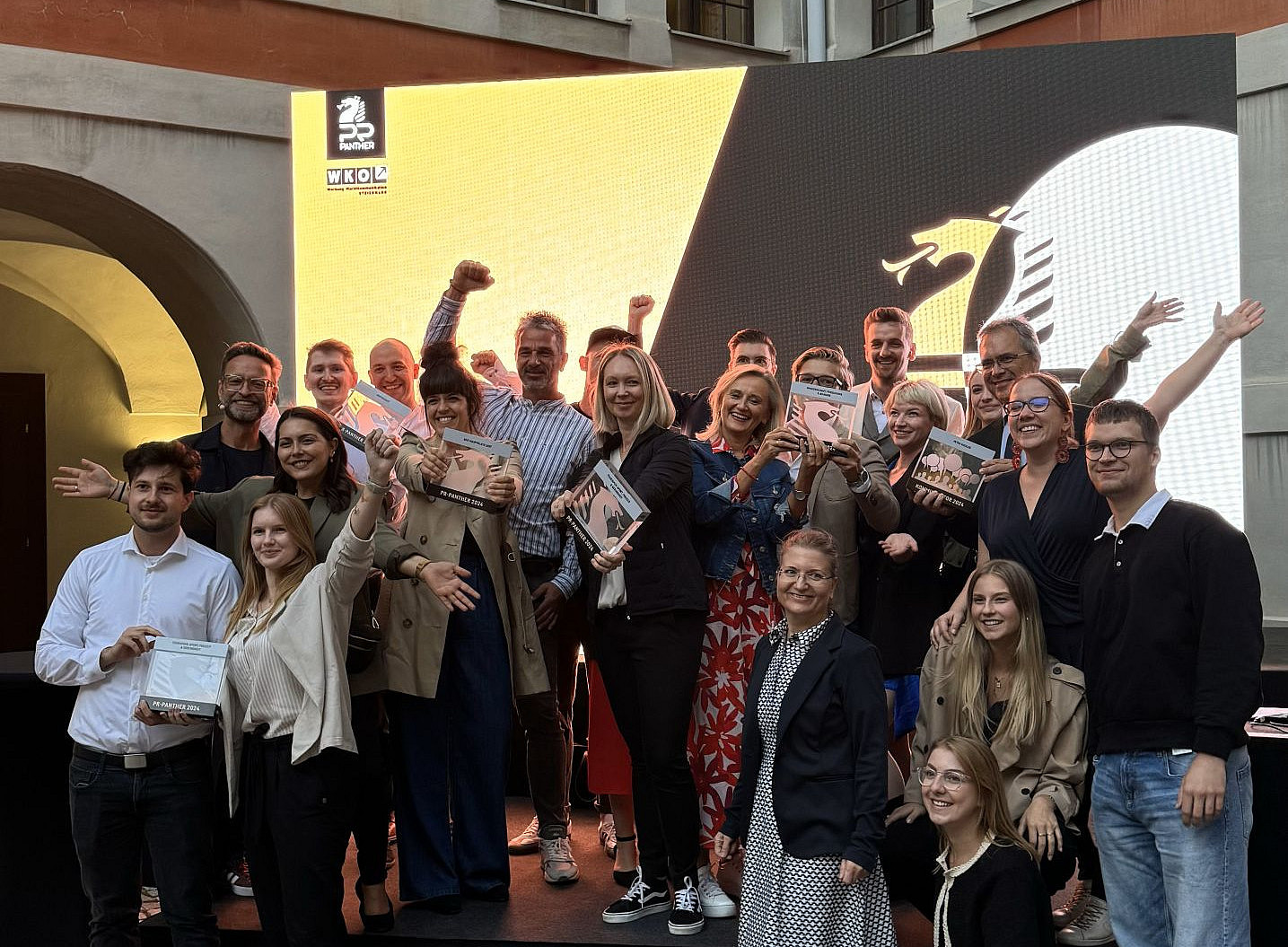 Ein Gruppenfoto von glücklichen Menschen, die die Preisverleihung für eine Werbekampagne feiern, vor einer schwarzen Werbetafel mit weißer Schrift und Markenzeichen darauf. Der Ort ist das Priesterseminar Graz. Es gibt Steinmauern und beigefarbene Säulen. 