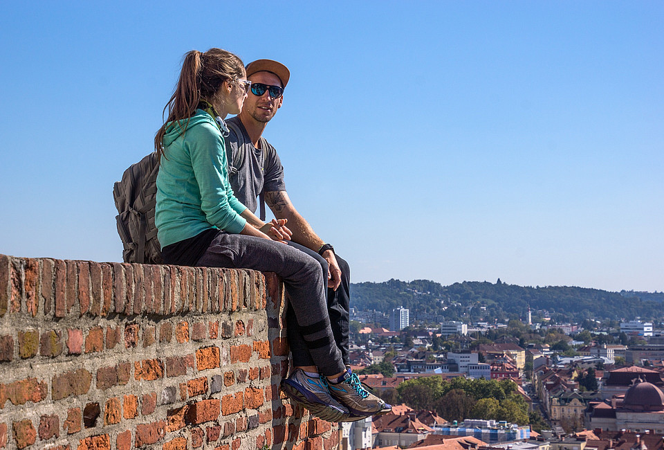 Eine Frau und ein Mann sitzen auf einer Ziegelmauer und schauen in die Ferne, im Hintergrund zeigt sich die Stadt Graz