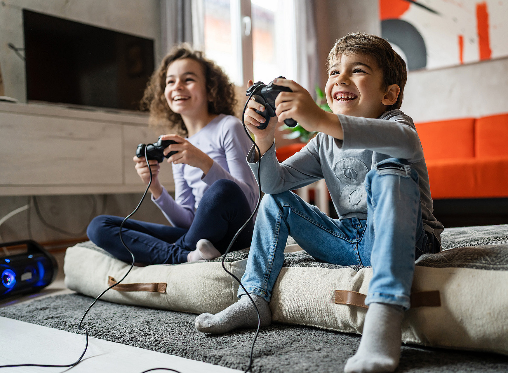 Ein Mädchen und ein Bub sitzen in einem Wohnzimmer auf dem Boden mit einem Controller in der Hand und spielen fröhlich ein Computerspiel. ©Miljan Zivkovic