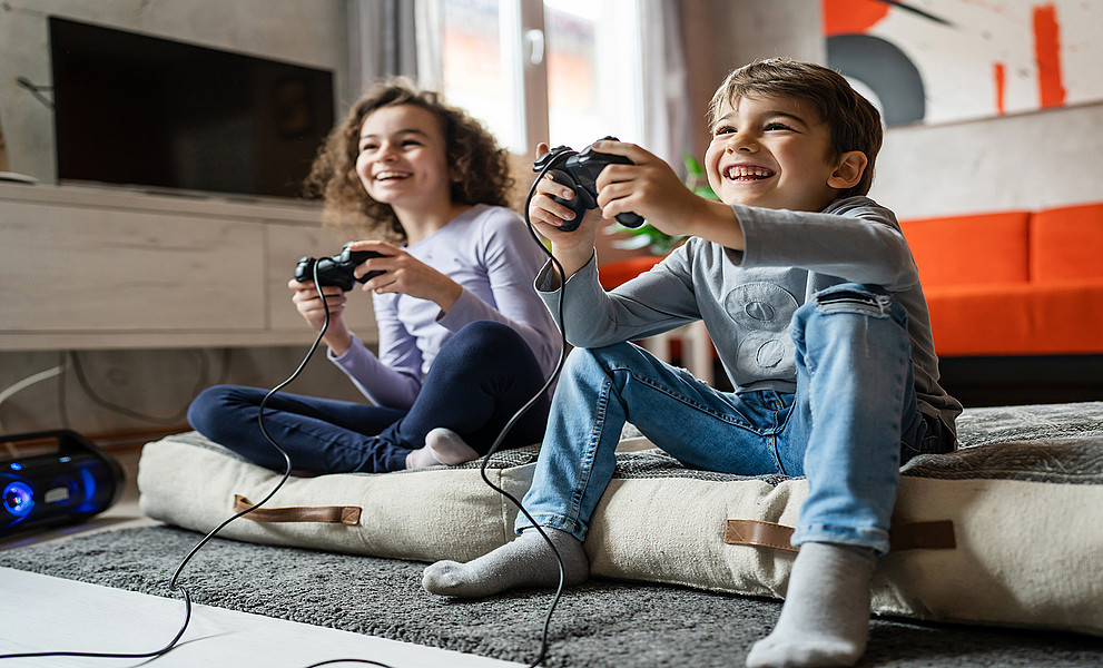 Ein Mädchen und ein Bub sitzen in einem Wohnzimmer auf dem Boden mit einem Controller in der Hand und spielen fröhlich ein Computerspiel.