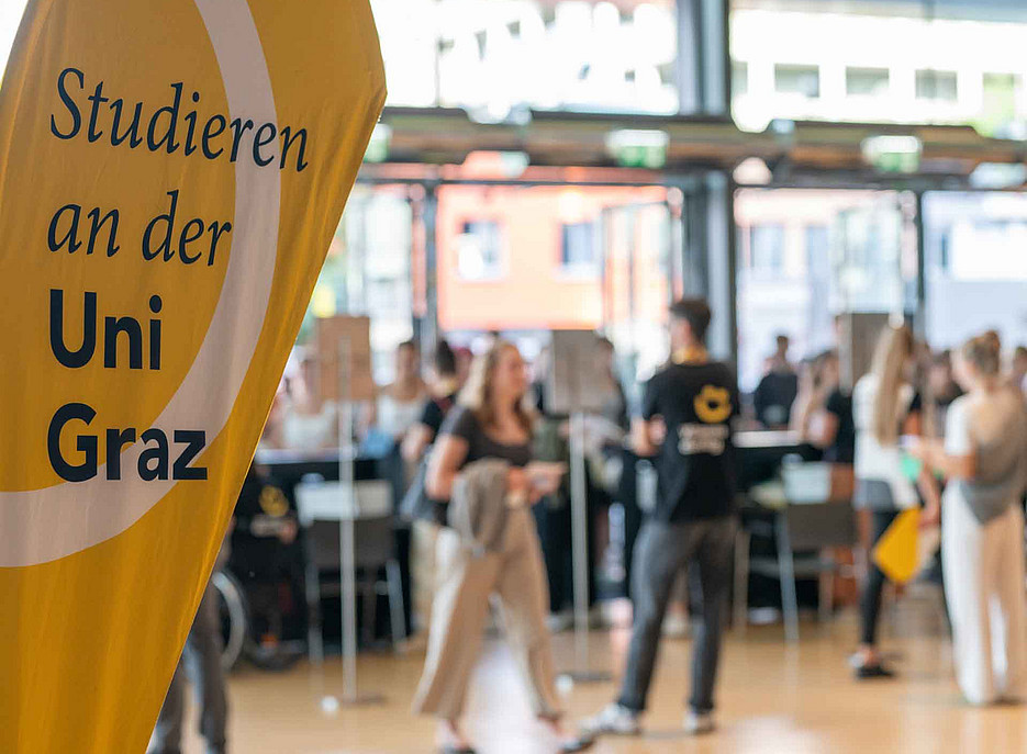 Gelbes Banner mit der Aufschrift „Studieren an der Uni Graz“, im Hintergrund mehrere junge Erwachsene in legerer Kleidung in einer hellen Halle. ©Uni Graz/Angele