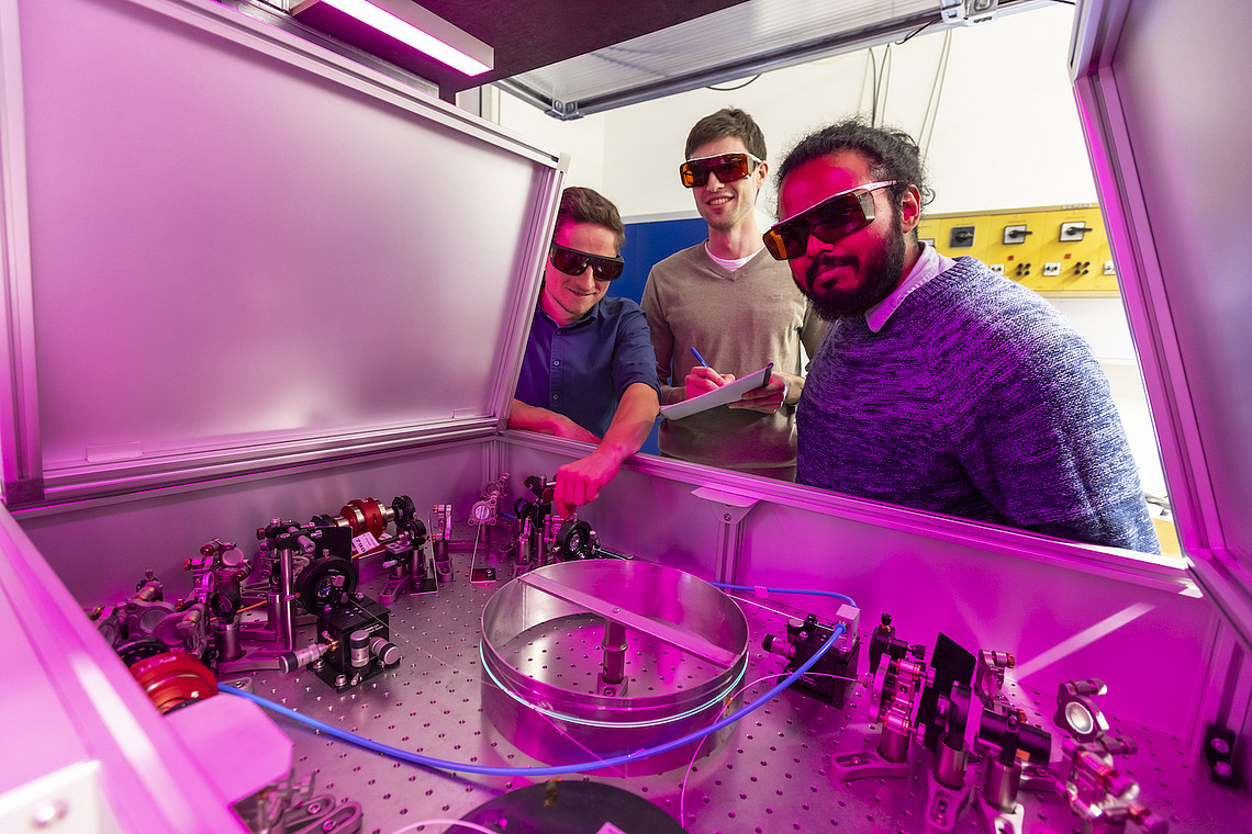 People wearing safety goggles stand in front of an open machine with a violet light.