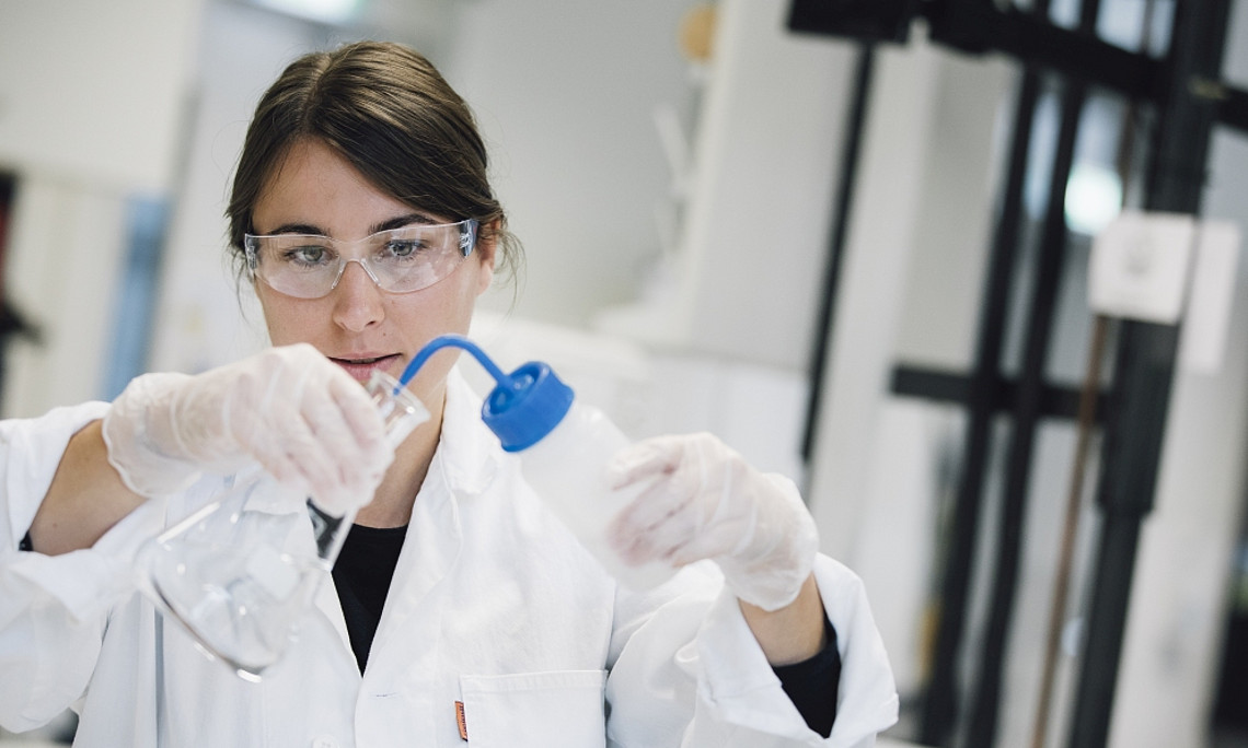 Eine Frau hält eine Pipette in ein Glas, trägt Laborkleidung und hat eine Schutzbrille auf