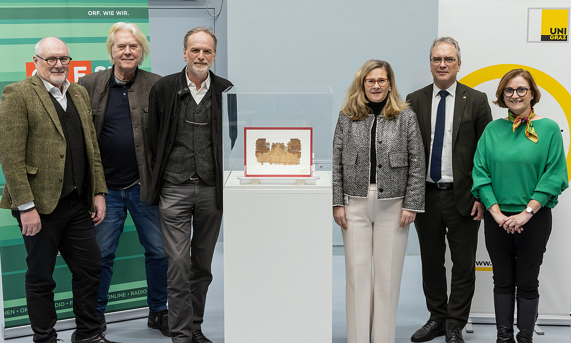 Ein Gruppenfoto an der Universität Graz, sechs Personen stehen nebeneinander, in der vorderen Mitte befindet sich ein altes Papyrusobjekt im Glas
