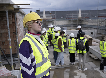 Weitwinkelaufnahme von jungen Menschen in Warnwesten und Schutzhelmen, die auf dem Dach einer aktiven Baustelle stehen, im Hintergrund ein Gebäude. Eine Frau blickt in die Kamera und lächelt; sie hat blonde Haare und eine Brille. ©Uni Graz/Jürgen Fuchs