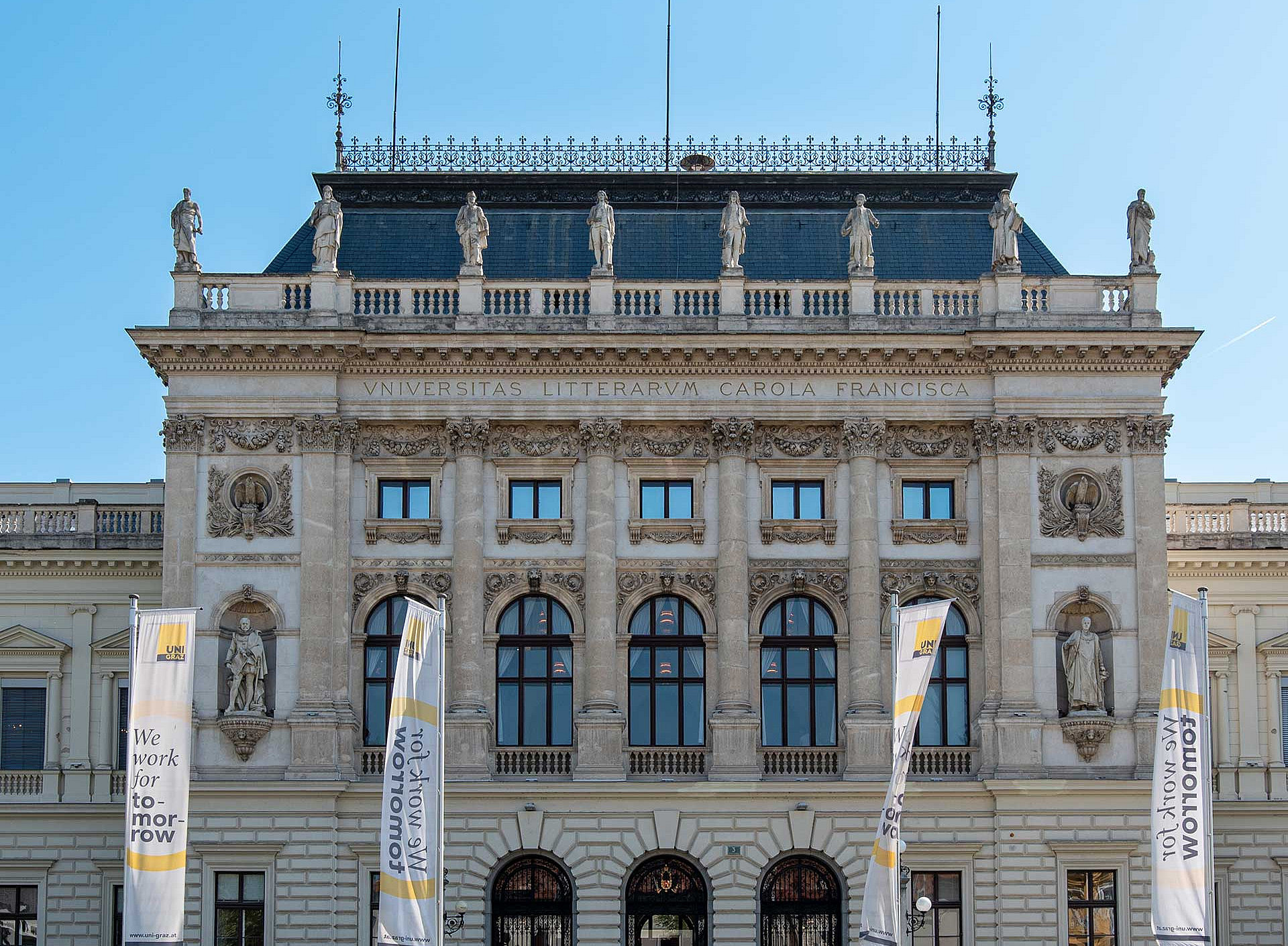Main building of the University of Graz with four ‘We work for tomorrow’ flags in front of it ©Universität Graz