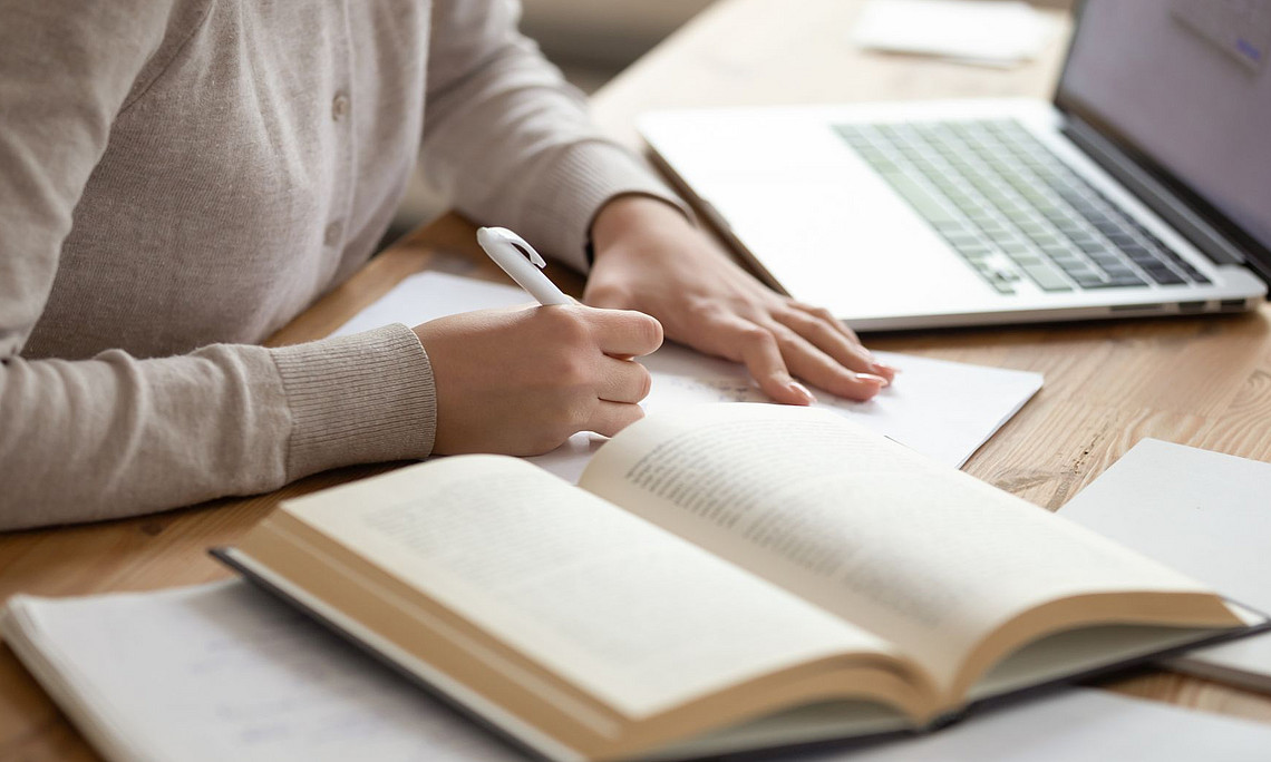 Person sitzt vor einem aufgeklappten Laptop und einem aufgeschlagenen Buch am Schreibtisch und arbeitet
