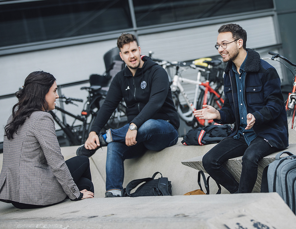Vier Studierende sitzen auf Betonbänken vor der Uni Graz und unterhalten sich.