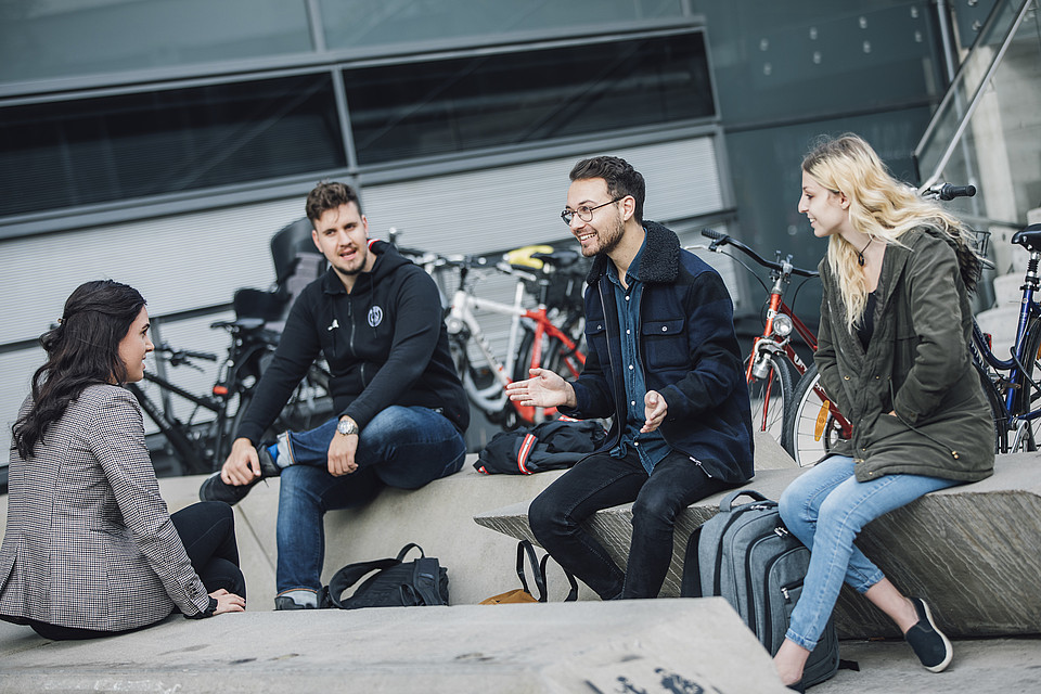 Vier Studierende sitzen auf Betonbänken vor der Uni Graz und unterhalten sich.