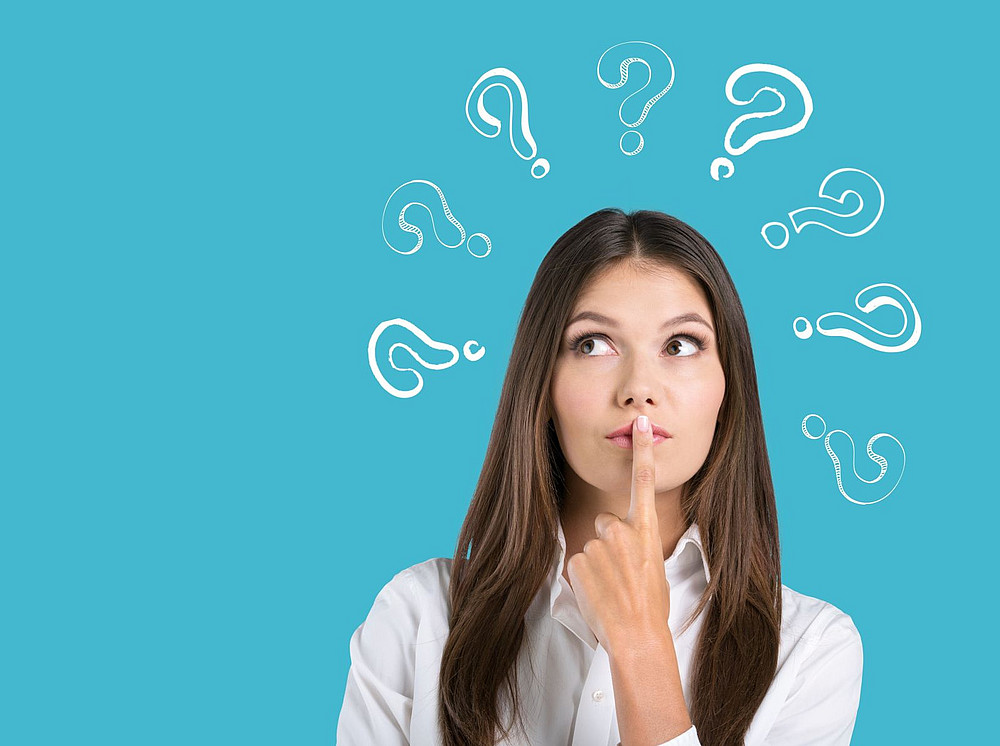 Young woman, white question marks appearing around her head against a blue background, holding her index finger to her lips. ©BillionPhotos.com - stock.adobe.com