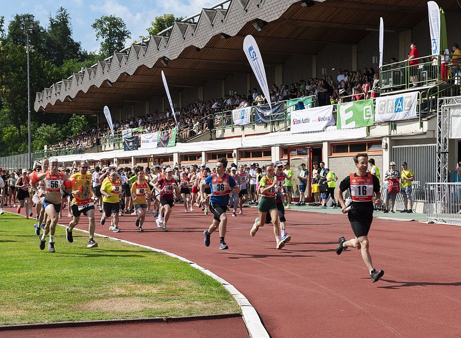 Zahlreiche Läuferinnen und Läufer mit Startnummern rennen auf einer roten Laufbahn in einem Stadion, während auf der Tribüne eines überdachten Gebäudes viele Zuschauer sitzen und bunte Fahnen sowie Werbebanner im Hintergrund zu sehen sind. ©Uni Graz/Angele