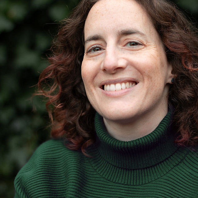 Ein Foto von Daniella Shaw, einer Frau mit lockigem braunem Haar, die lächelt und einen dunkelgrünen, gerippten Rollkragenpullover trägt. Sie steht im Freien vor einem Hintergrund aus grünen Blättern. ©Daniella Shaw