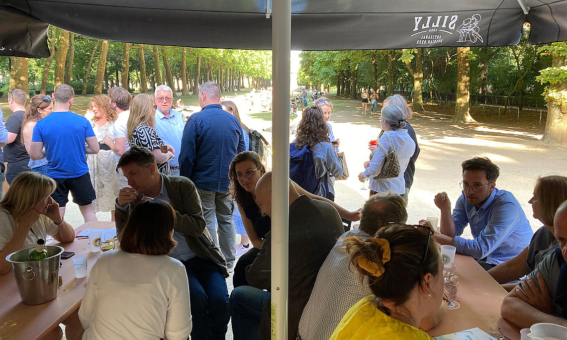 Personen die im Vordergrund auf Holztischen und Holzbänken sitzen, im Hintergrund stehen viele Personen, sie sind in Gespräche vertieft. Im Hintergrund lässt sich ein Park und Bäume erkennen.
