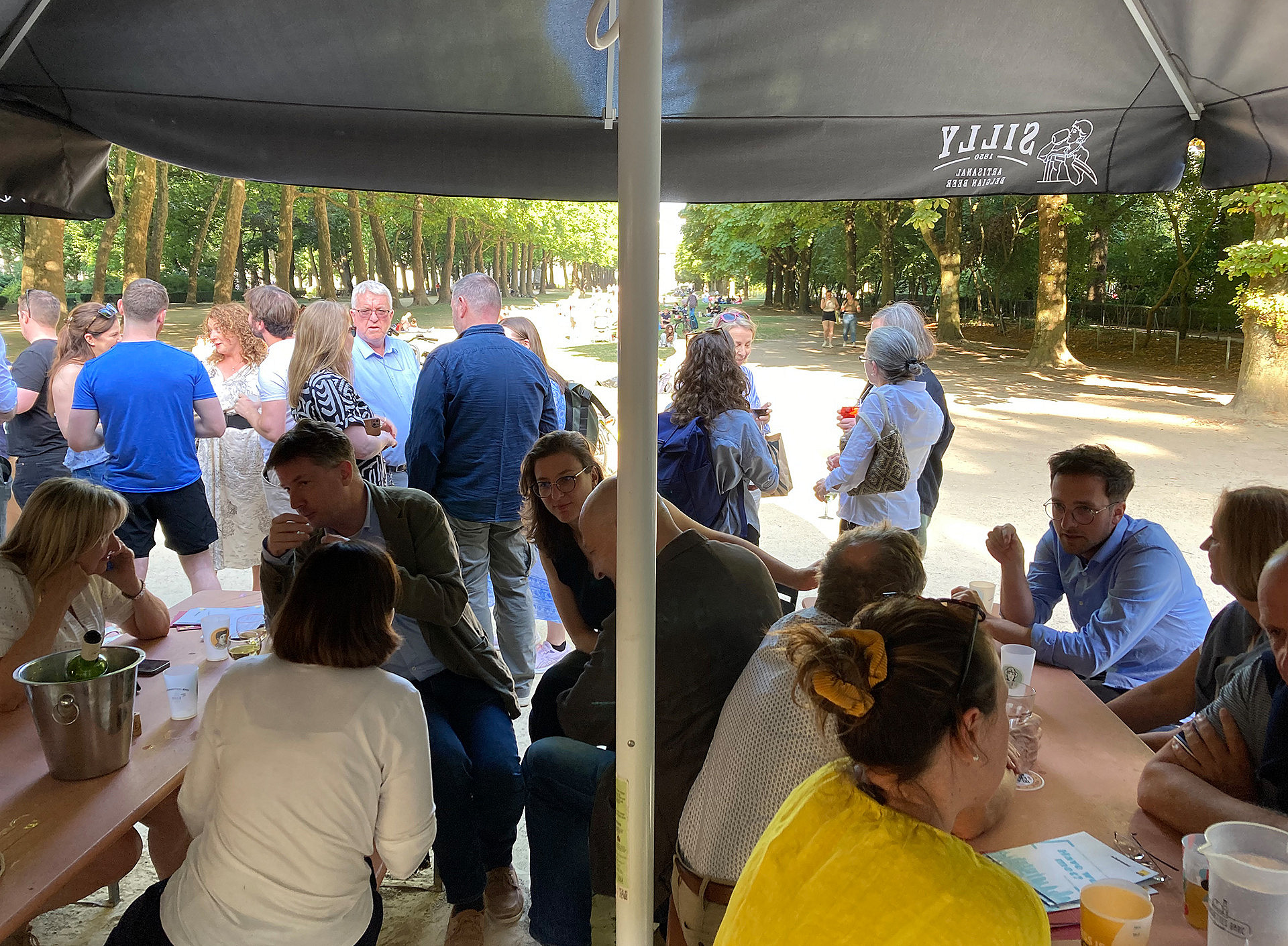 Personen die im Vordergrund auf Holztischen und Holzbänken sitzen, im Hintergrund stehen viele Personen, sie sind in Gespräche vertieft. Im Hintergrund lässt sich ein Park und Bäume erkennen. ©Verena Martelanz