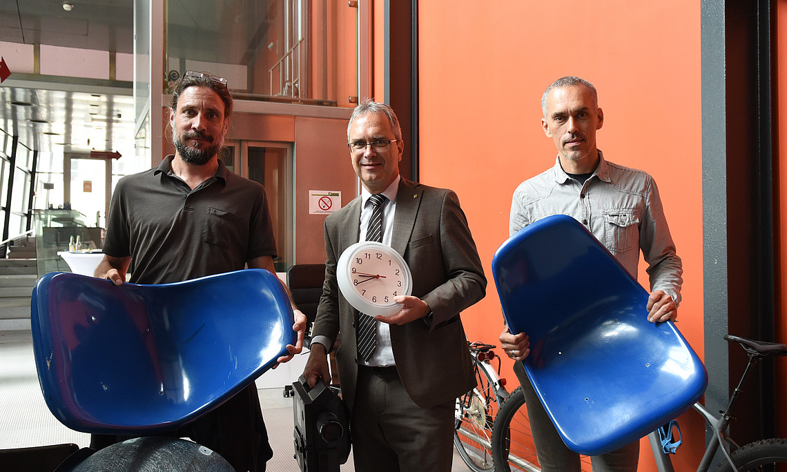 Philipp Jereb (Bundesimmobiliengesellschaft), Rektor Peter Riedler (Uni Graz) und Anton Edelsbrunner (Soziale Arbeit Steiermark) mit Hörsaal-Sesseln, einer Uhr und einem Diaprojektor in der Vorklinik. 