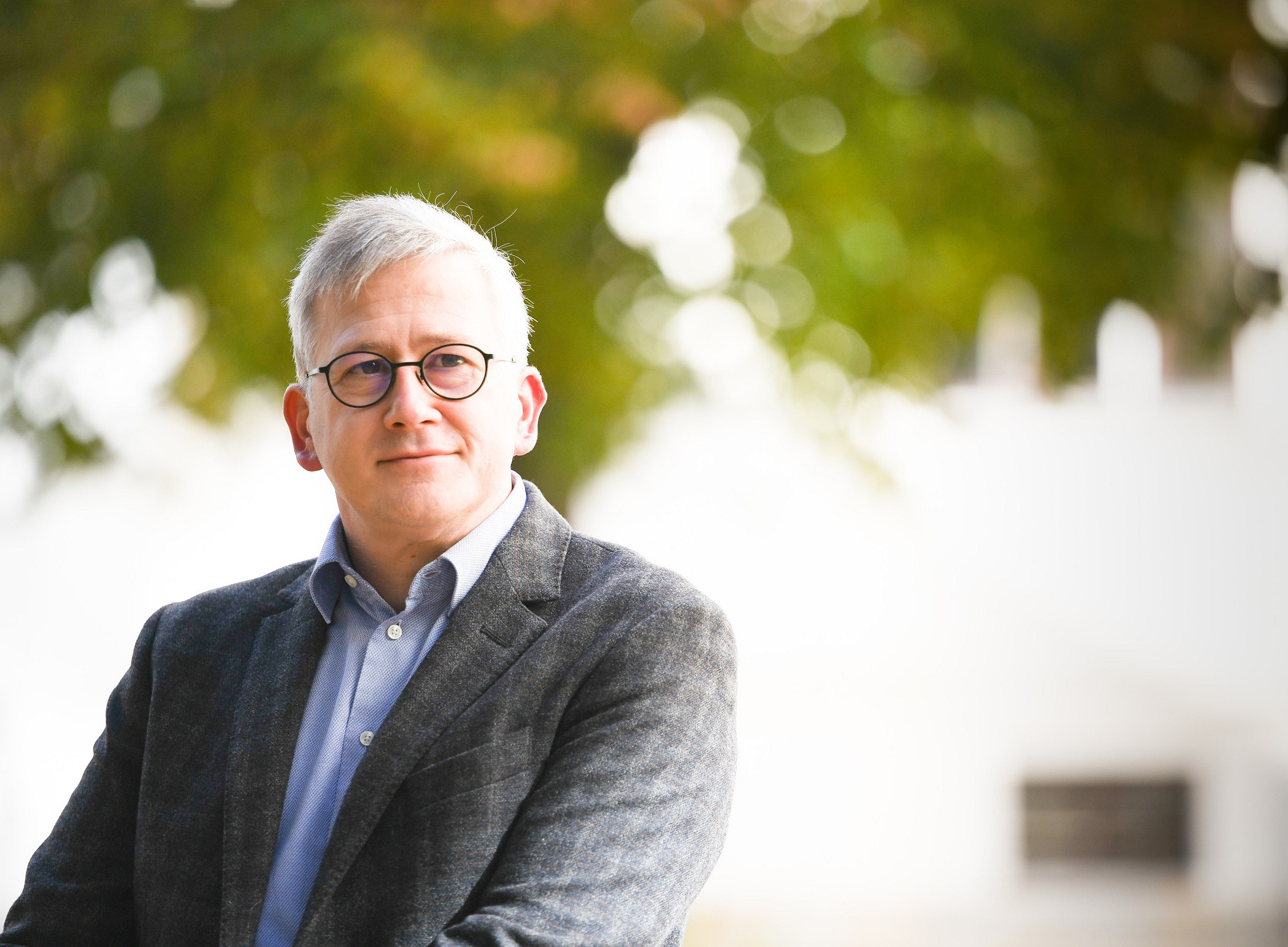 Ein Mann in einem grauen Sakko und hellblauem Hemd steht vor einem unscharfen Hintergrund mit grünen Bäumen und einem weißen Gebäude. ©Universität Graz/Tzivanopoulos