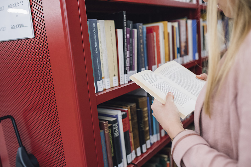 Nahaufnahme einer Studentin, die in einer Bibliothek vor einer Bücherwand in einem Buch liest