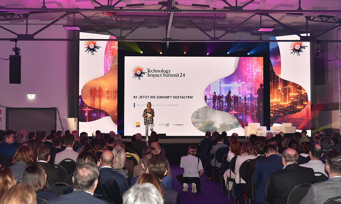 a large hall with audience, stage and big led screen displaying the logo of " technology impact summit" conference in white background. a woman is standing on the stage giving speech to crowd people sitting below her at a business event. 
