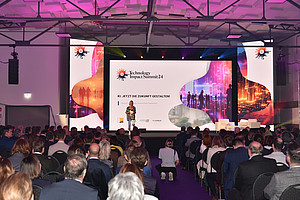 a large hall with audience, stage and big led screen displaying the logo of " technology impact summit" conference in white background. a woman is standing on the stage giving speech to crowd people sitting below her at a business event. 