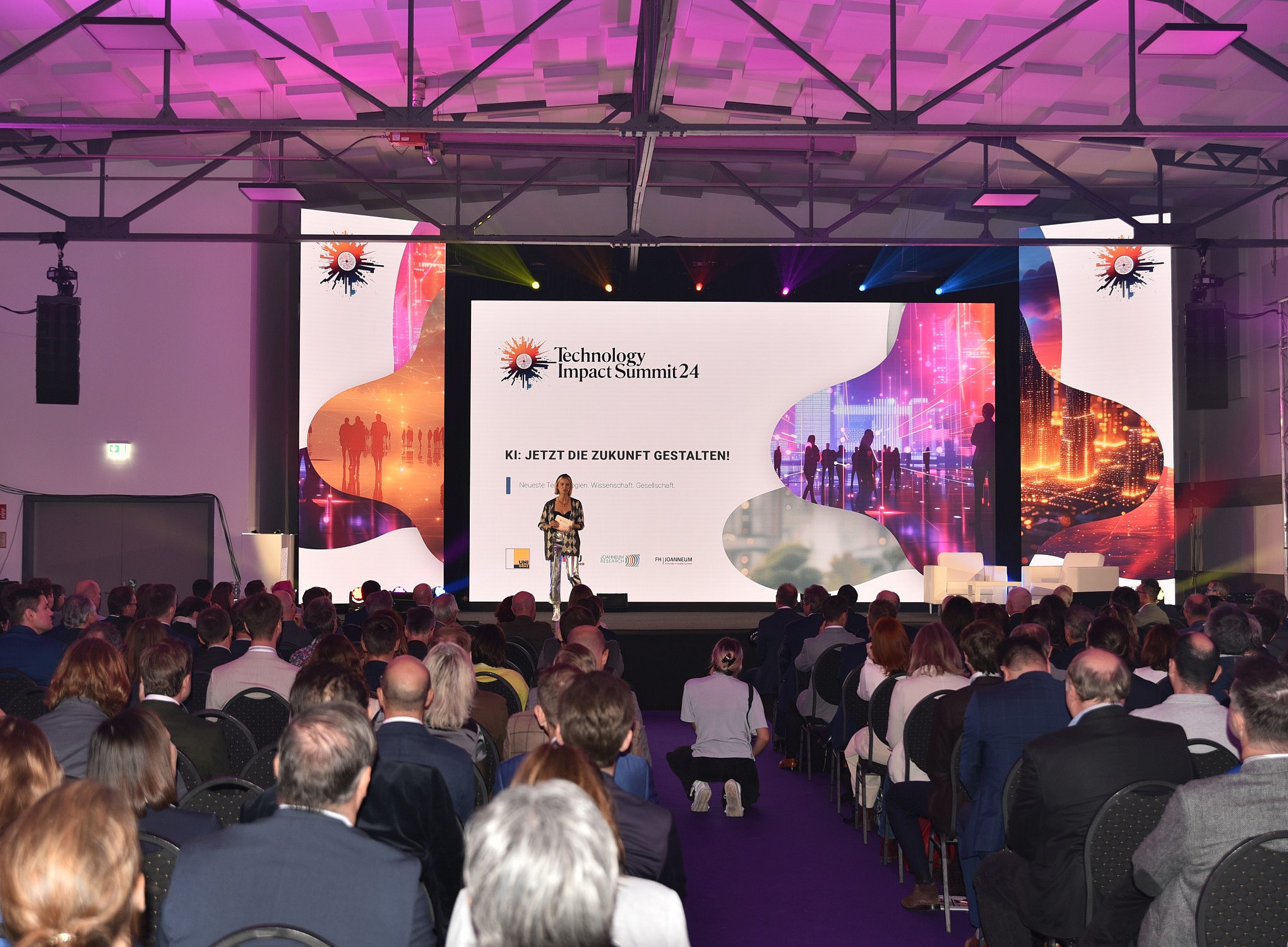 Ein großer Saal mit Publikum, Bühne und großem LED-Bildschirm, auf dem das Logo der Konferenz „Technology Impact Summit“ vor weißem Hintergrund zu sehen ist. Eine Frau steht auf der Bühne und hält vor den unter ihr sitzenden Menschen eine Rede bei einer Geschäftsveranstaltung. 