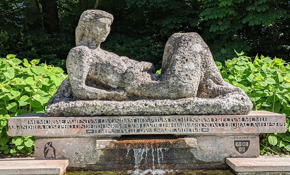 Steinfigur liegt auf einem Podest unterhalb eine lateinische Inschrift