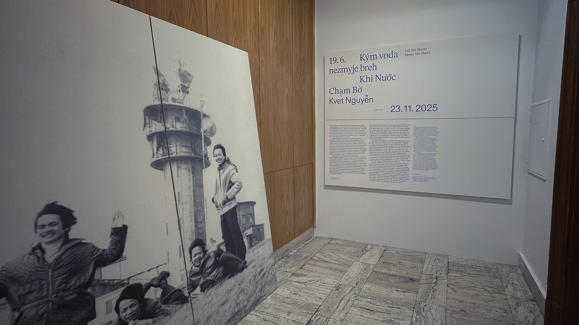 Gallery corner with a large black-and-white photographic installation leaning against a wood-paneled wall. The photo shows several smiling people posing outdoors near a tall lighthouse-like structure. On the adjacent white wall, an exhibition panel displays the title “Kým voda nezmyje breh / Khi Nước Chạm Bờ” by Kvet Nguyen with the date 23.11.2025, along with blocks of explanatory text. The floor is tiled in stone, and the space appears quiet and minimal. ©Stefanie Populorum