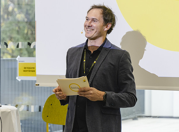 Ein lächelnder Mann mit Headset hält bei einer Veranstaltung eine Karte mit der Aufschrift „We work for tomorrow“. ©@foto-gasser.at