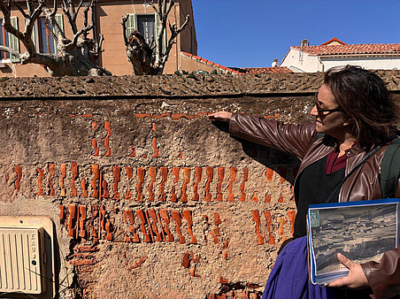 A person standing beside a weathered wall with exposed brick patterns, pointing to its surface while holding an old photograph, with houses and leafless trees in the background under a clear sky. ©Ben Gidley