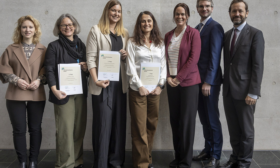 A group of seven people are standing side by side in front of a gray wall. Three people in the middle are holding certificates in their hands. The people are mostly dressed formally, in blazers, shirts, and suit pants. The floor is covered with dark tiles.
