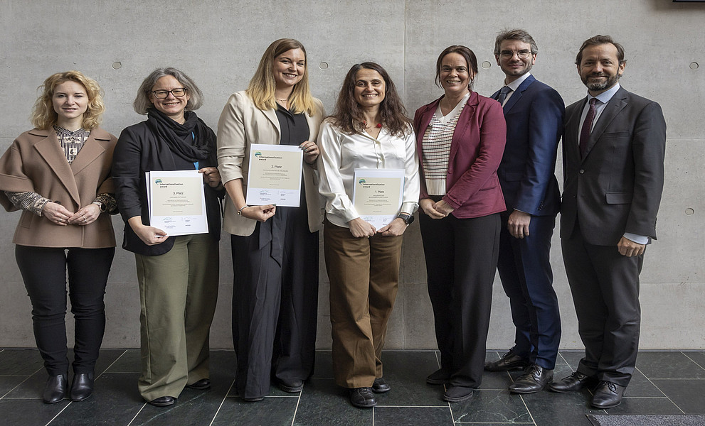 Eine Gruppe von sieben Personen steht nebeneinander vor einer grauen Wand. Drei Personen in der Mitte halten Zertifikate in den Händen. Die Kleidung der Personen ist überwiegend formal, mit Blazern, Hemden und Anzughosen. Der Boden besteht aus dunklen Fliesen.