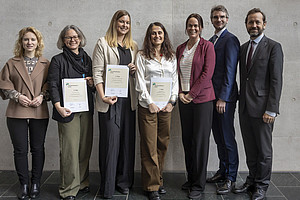 A group of seven people are standing side by side in front of a gray wall. Three people in the middle are holding certificates in their hands. The people are mostly dressed formally, in blazers, shirts, and suit pants. The floor is covered with dark tiles.