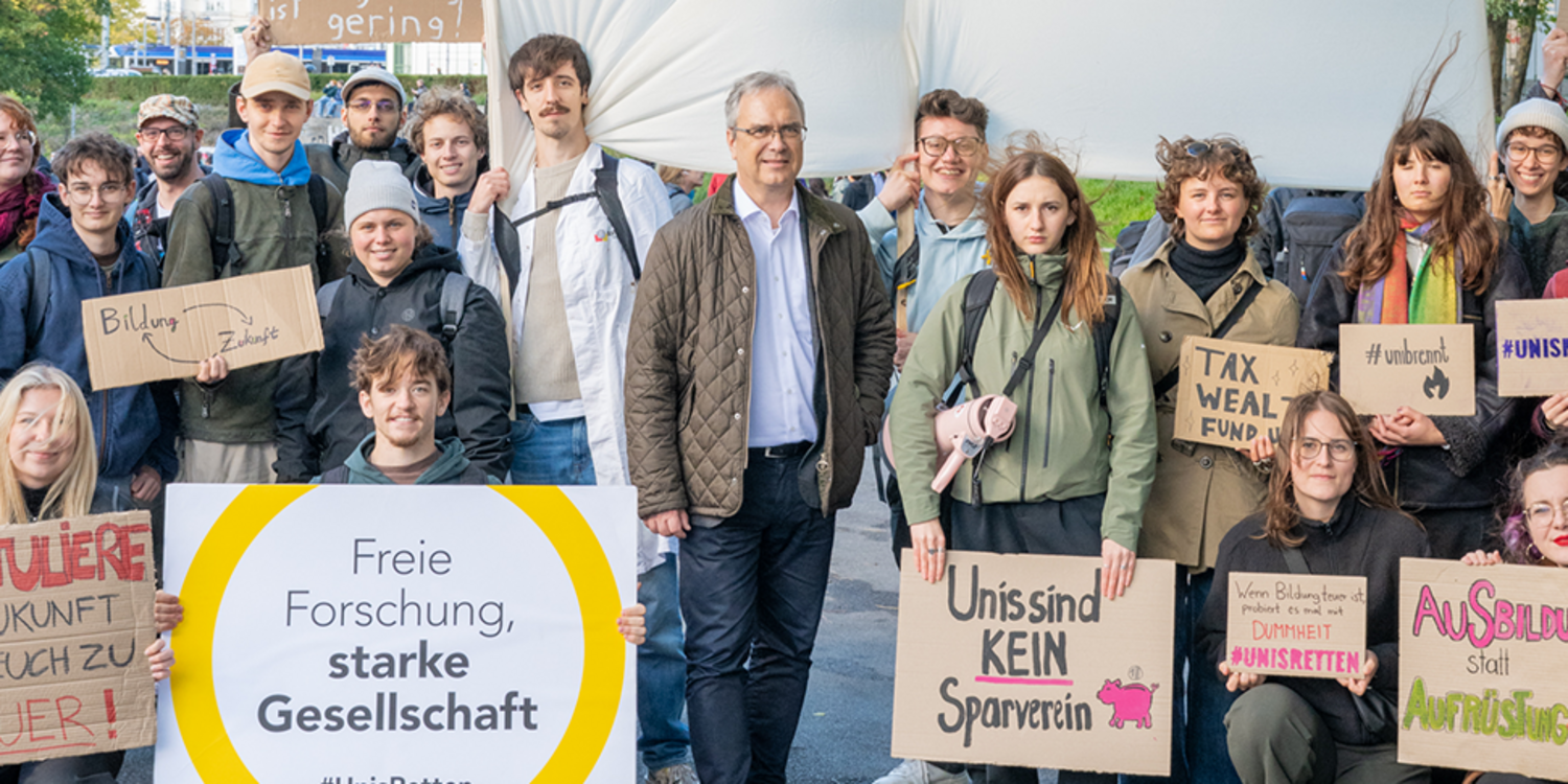 Gruppe überwiegend junger Menschen steht im Freien und hält Protestbanner und Schilder gegen Budgetkürzungen an Universitäten hoch.