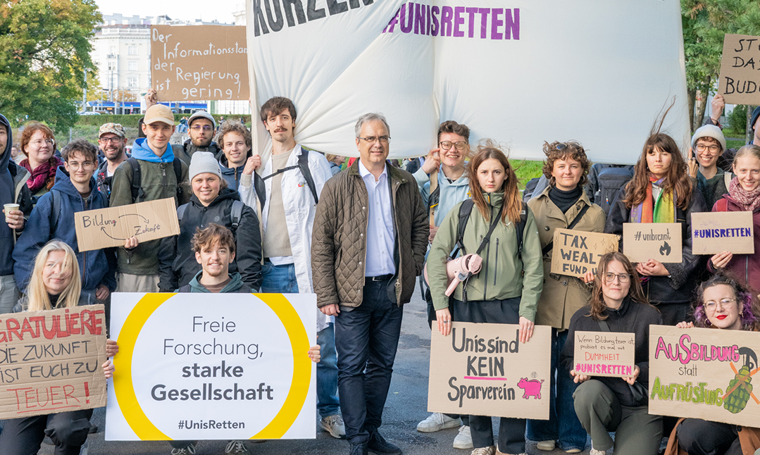 A group of mostly young people are standing outside, holding up protest banners and placards against budget cuts at universities. 