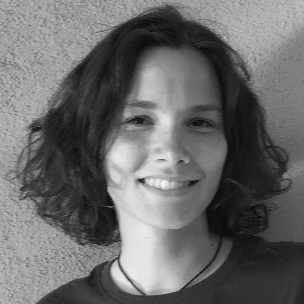 Ein Schwarz-Weiß-Porträt einer lächelnden Person mit kurzen, leicht gewellten Haaren, die bis etwa zum Kinn reichen. Die Person blickt direkt in die Kamera. Ihr Gesicht ist gleichmäßig ausgeleuchtet, und sie trägt eine schlichte Halskette mit einem dünnen Band. Der Hintergrund besteht aus einer strukturierten, hellen Wand. ©Giulia Buffoli