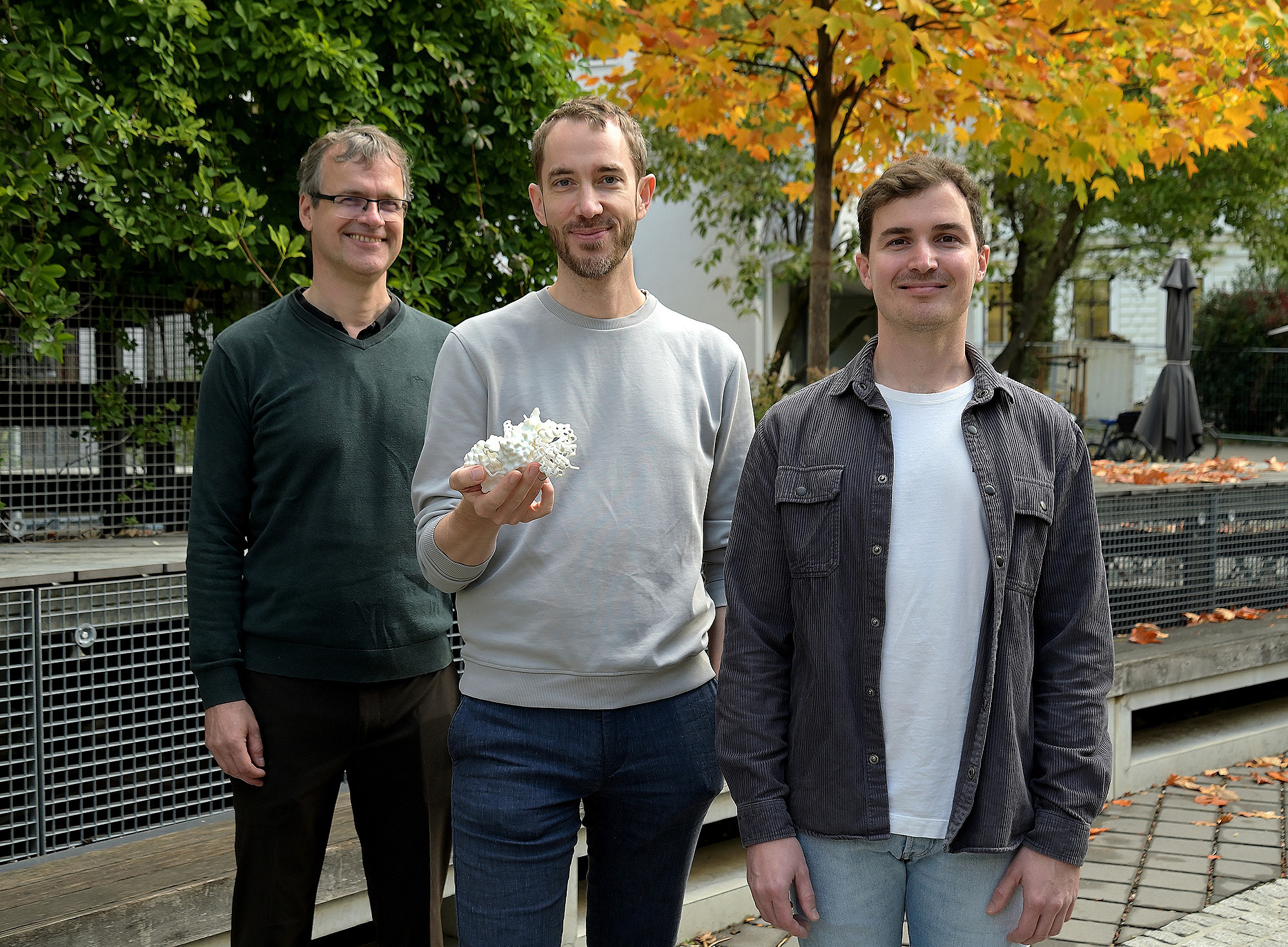 Drei Männer mittleren Alters stehen draußen vor Herbstbäumen; der mittlere hält ein weißes Modell eines Enzyms in der Hand. ©Uni Graz/Pichler