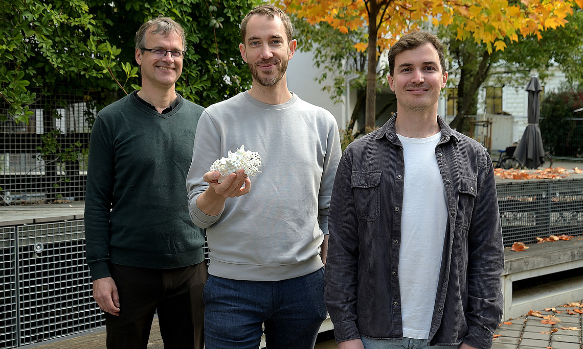 Drei Männer mittleren Alters stehen draußen vor Herbstbäumen; der mittlere hält ein weißes Modell eines Enzyms in der Hand.