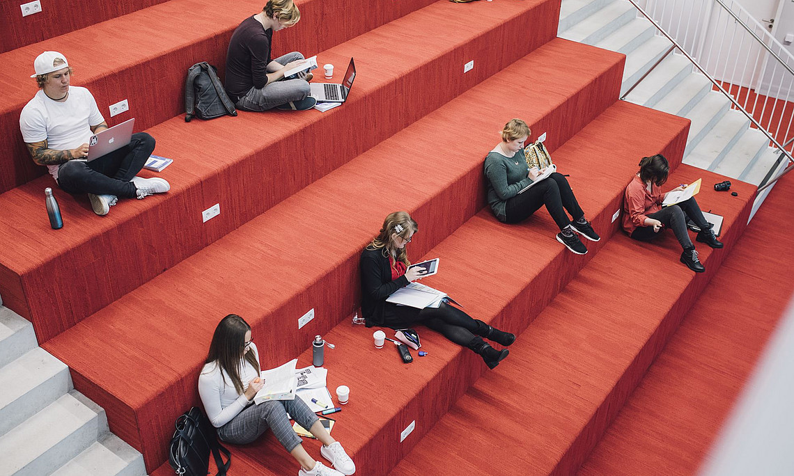 A group of students are sitting on a red staircase in the interior, studying and working on laptops.