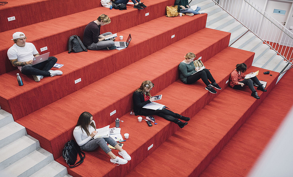Eine Gruppe von Studenten sitzt auf einer roten Treppe im Innenraum und lernt und arbeitet mit Laptops.