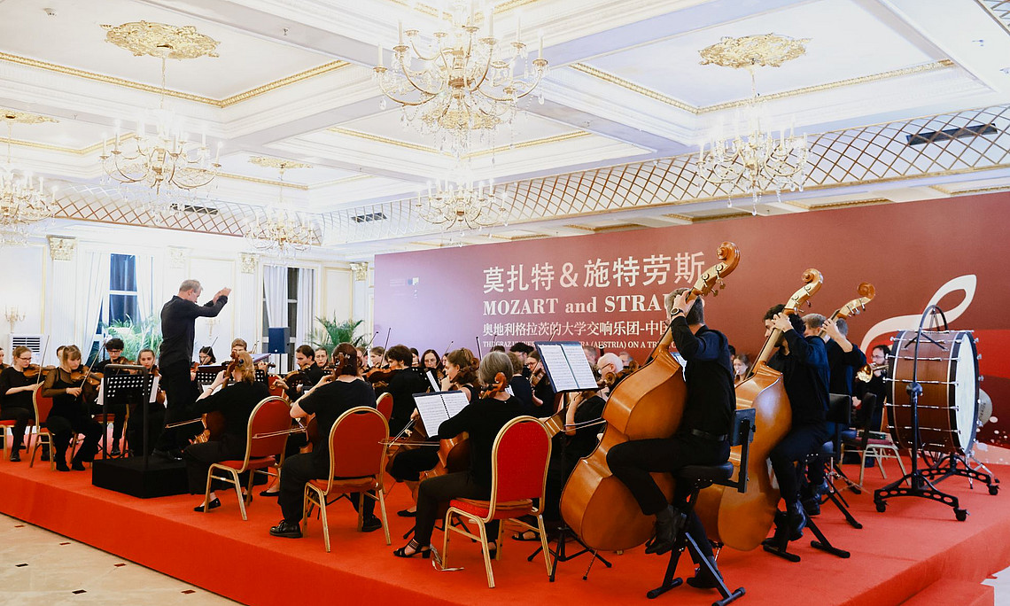  A photo of an orchestra playing on a stage, and musical instruments such as cellos, drums, flutes, chairs arranged for audience seats, a red carpet leading to the scene from one side, all set against luxurious wall hangings and chandeliers. The background is decorated with large Chinese-style banners showing a performance called "Mozart & Strauss"