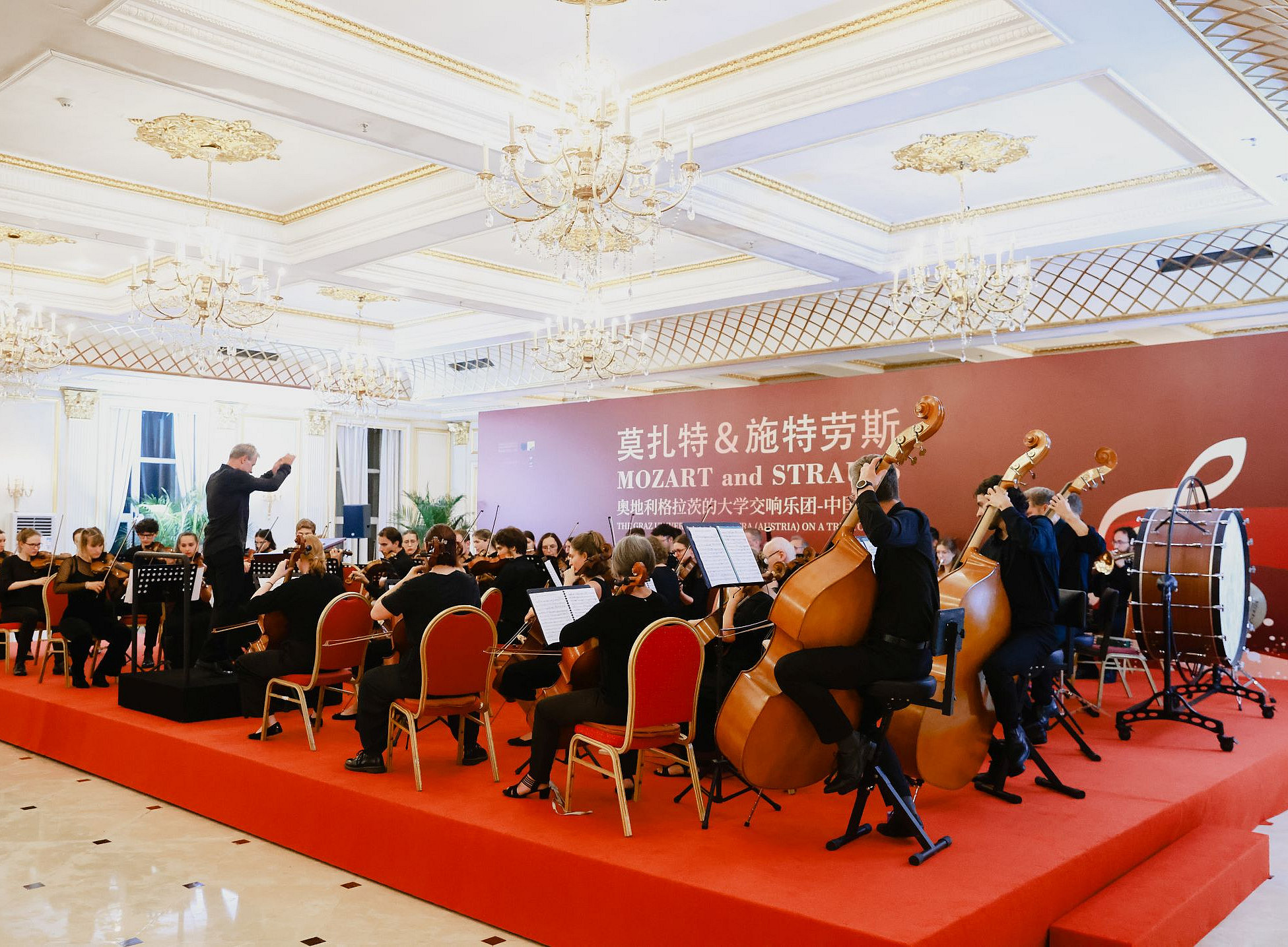 Ein Foto eines Orchesters, das auf einer Bühne spielt, und Musikinstrumente wie Celli, Trommeln, Flöten, Stühle, die als Zuschauerplätze angeordnet sind, ein roter Teppich, der von einer Seite zur Szene führt, alles vor luxuriösen Wandbehängen und Kronleuchtern. Der Hintergrund ist mit großen Bannern im chinesischen Stil geschmückt, die eine Aufführung mit dem Titel „Mozart & Strauss“ zeigen. 