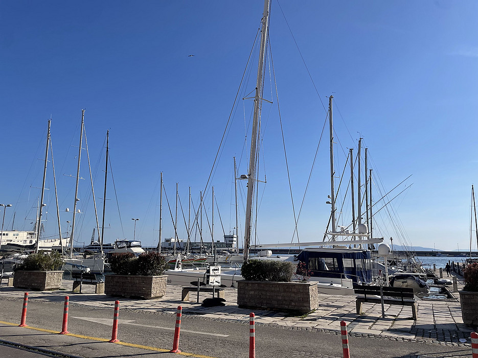 A marina under a clear blue sky with several sailboats docked. Tall masts create a vertical pattern. Potted plants and bollards line the walkway. ©Milka Car