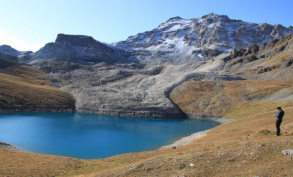Blockgletscher, der in einen Gebirgssee mündet, schneebedeckter Fels im Hintergrund