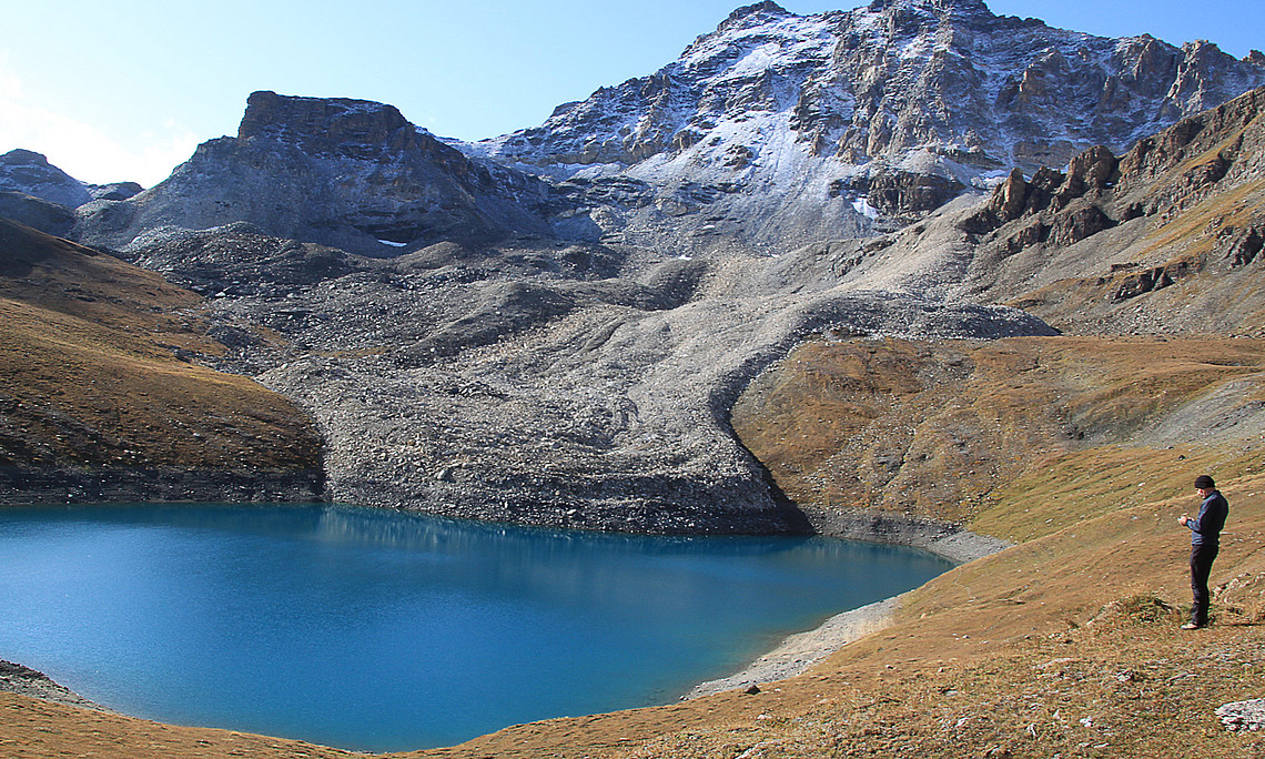 Blockgletscher, der in einen Gebirgssee mündet, schneebedeckter Fels im Hintergrund